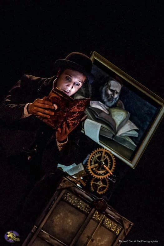 Charly Brahim, chapeau melon, lisant un grimoire éclairé sur scène devant un tableau et des rouages d’horlogerie dans Le Maître du Temps